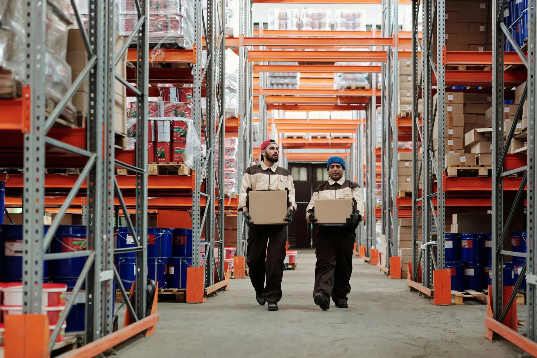 Two warehouse workers carrying boxes down an aisle of tall metal shelves